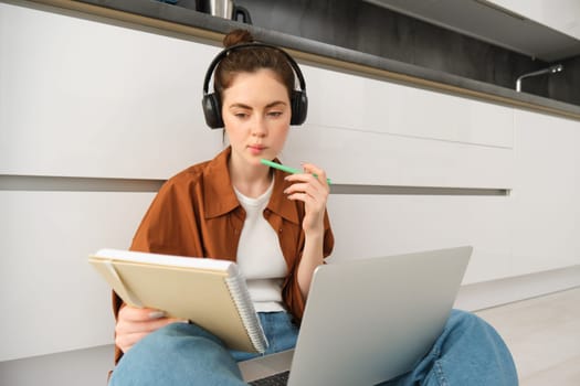 Serious-looking girl student studying at home, sits with laptop and notebook on floor, doing homework project on computer, has online course lesson, wears headphones.