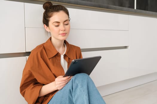 Portrait of cute young woman, freelancer working from home, sitting on kitchen floor with digital tablet and smiling.