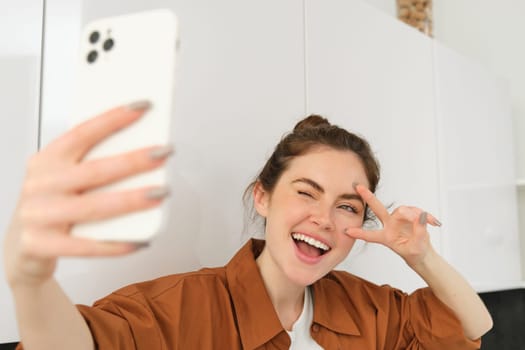 Close up portrait of positive, happy young woman taking selfie on mobile phone, posing for photo on smartphone, showing peace sign and smiling at telephone camera.