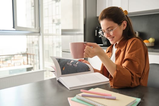 Portrait of woman reading at home, flipping pages of favourite book, relaxing in kitchen with cup of coffee, wearing glasses.