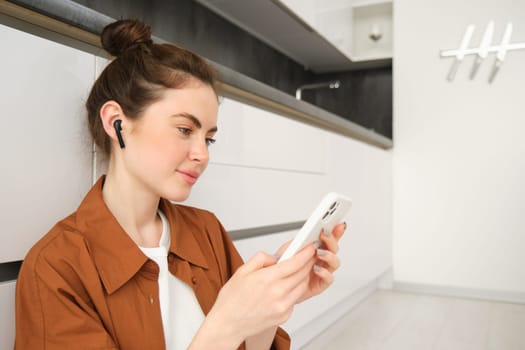 Close up portrait of young modern woman sits on kitchen floor, looking at her smartphone, watching video or tv series on streaming service app, listening music in wireless headphones.