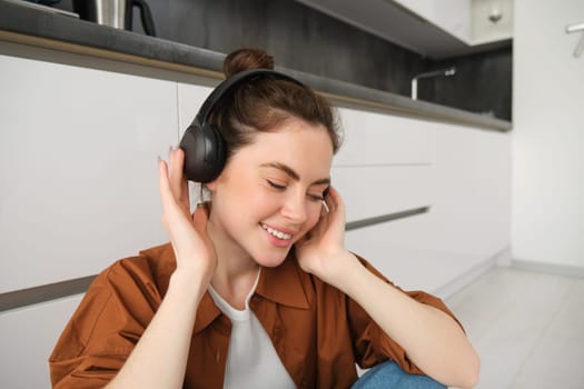 Cute brunette woman on kitchen floor, wears headphones, listens music and smiles, enjoys sound quality.