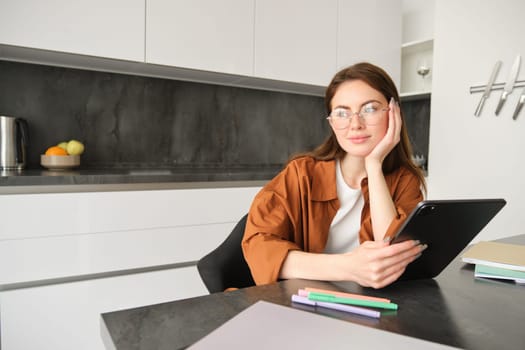 Portrait of young student, woman studying at home, working remotely, setup workplace in her kitchen, sitting on chair with digital tablet, reading in glasses.