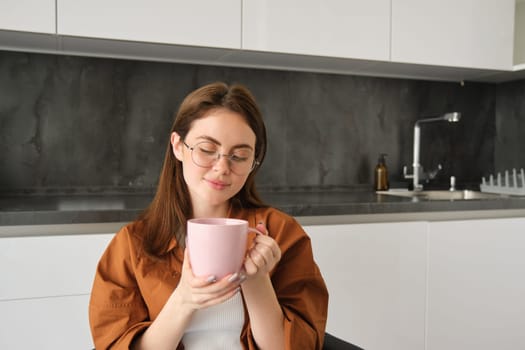 Relaxing day at home. Smiling young woman in glasses, enjoying fresh brewed cup of coffee, holding mug and sitting in kitchen, resting on peaceful autumn day at home.