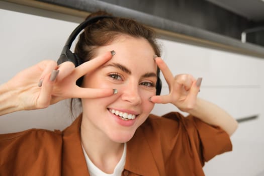 Close up of carefree young woman, laughing and smiling, showing peace, v-sign gesture, listening to music in wireless headphones.