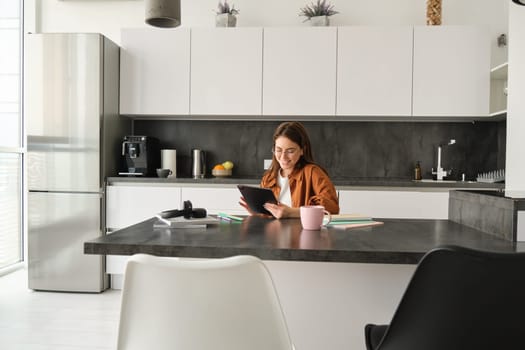 Portrait of young woman, student sitting in kitchen with tablet and notebook, reading news online, studying remotely, e-learning at home.