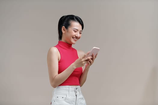 Beautiful young asian woman holding smartphone standing on isolated beige background.