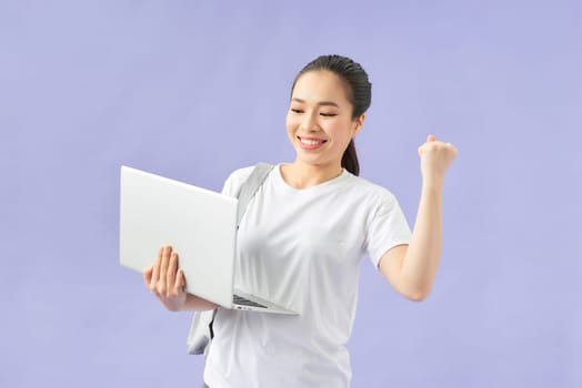 joyful asian student with backpack holding laptop isolated on violet
