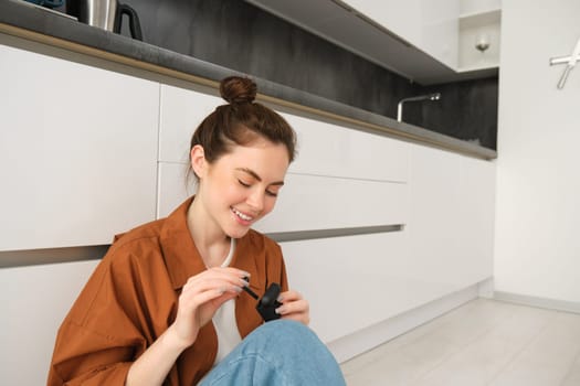 Portrait of beautiful woman enjoys listening to music or podcast in wireless black headphones, using earphones, smiling and looking happy, sitting at home on kitchen floor.