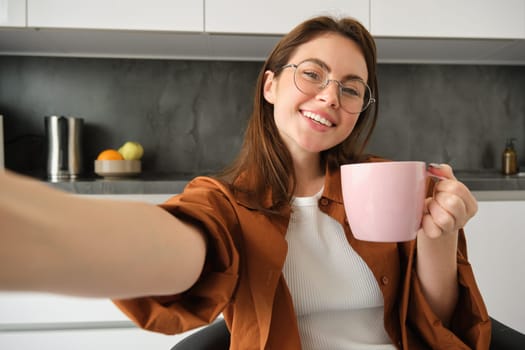 Portrait of happy young woman in glasses, takes selfie with smartphone, drinks coffee, posing with delicious mug of tea in kitchen.