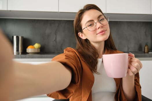 Portrait of beautiful young woman in glasses, takes selfie with cup of tea, sitting in kitchen, holding smartphone.