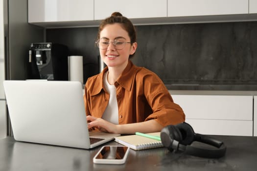 Portrait of smiling cute woman in kitchen, freelancer working from home on laptop, digital nomad freelancing from home, typing on computer.