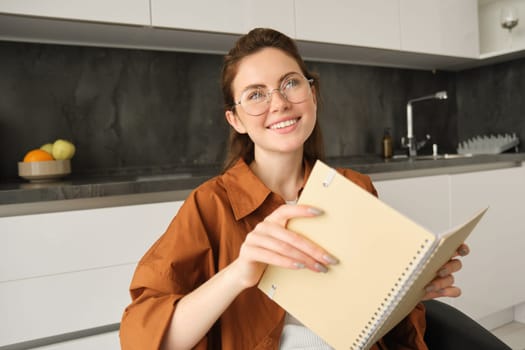 Portrait of beautiful young woman studying, student revising at home, doing homework, holding planner, reading her notes and smiling, writing in diary.