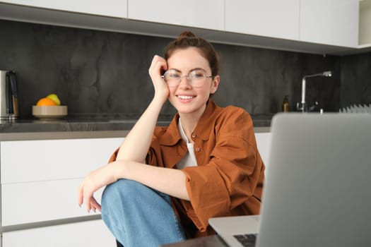 Portrait of young woman, business owner working from home, freelancer using laptop, sitting in kitchen, wearing glasses.