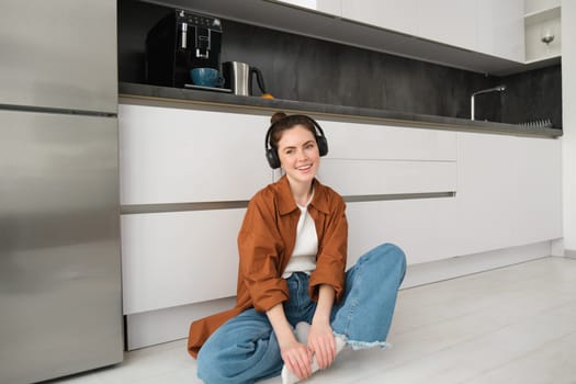 Cute brunette woman on kitchen floor, wears headphones, listens music and smiles, enjoys sound quality.