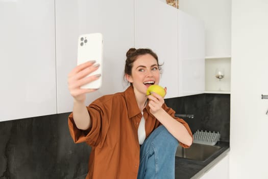 Happy woman using smartphone to take selfie, eating an apple on camera, records video for social media application, sitting on kitchen counter.