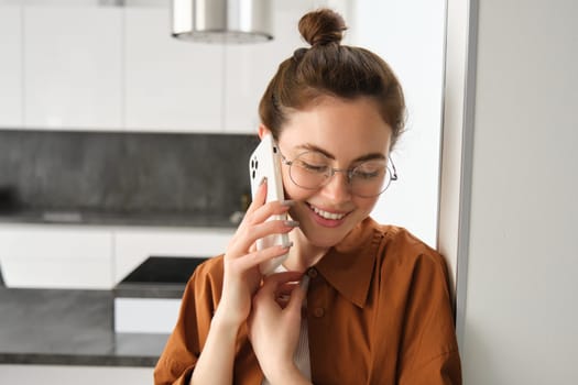 Portrait of lovely young woman answers phone call and smiling, leaning on wall, standing at home and talking on telephone.