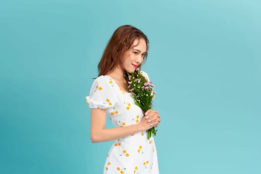 Image of happy woman in elegant dress and straw hat holding flowers isolated over blue background