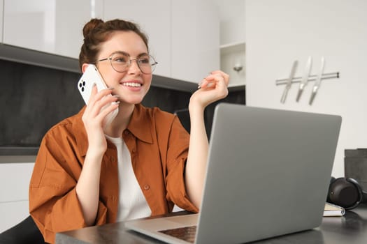 Portrait of young self-employed woman, entrepreneur working from home, freelancer calling client. Girl making an order, talking to someone on phone, sitting in kitchen with laptop.