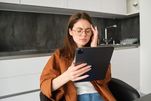 Portrait of woman freelancer, wearing glasses, holding digital tablet, looking tired, has migraine or headache, reading e-book. Freelance worker sits at home with gadget.