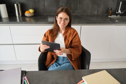 Portrait of young self-employed woman sitting at home in kitchen, reading papers, holding digital tablet, wearing glasses.