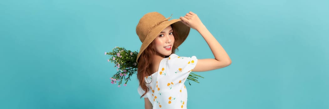 Image of happy woman in elegant dress and straw hat holding flowers isolated over blue background