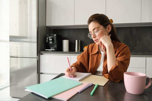 Portrait of young woman student, girl studying at home, working remote in kitchen, taking notes, sitting indoors.