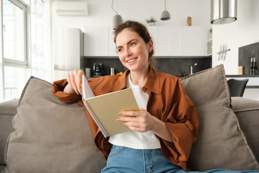 Smiling young brunette woman, sits on sofa in living room, holds notebook, reads her notes, studies for exam, student does her homework at home.