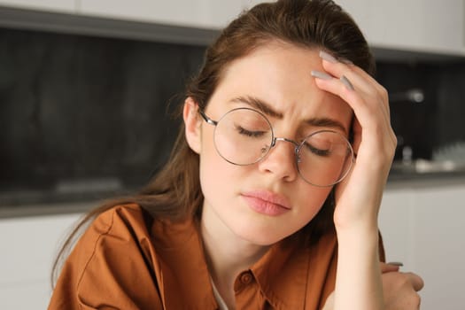 Portrait of tired young working woman in glasses, holds hands on head, suffers painful migraine, has pain in temples, sitting with headache in kitchen.