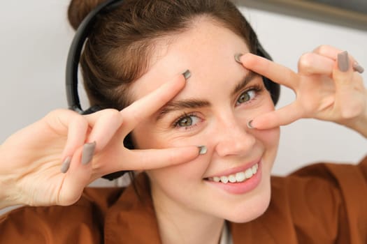 Close up portrait of beauty girl, listens music in wireless headphones, shows peace signs near face and smiles at camera.