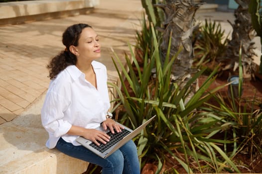 Beautiful mixed race young adult female student in white shirt and casual denim, remotely studying on laptop outdoor. People. Online education. Internet business. People. Career. Recruitment