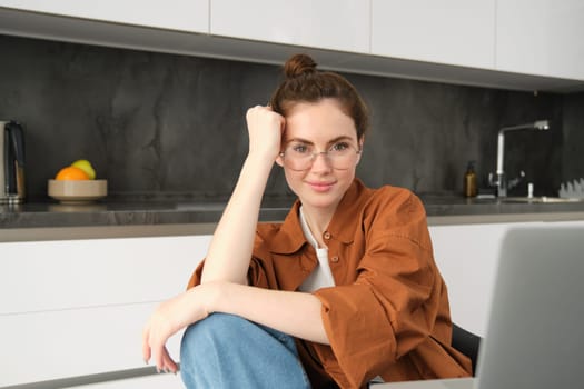 Portrait of young woman, business owner working from home, freelancer using laptop, sitting in kitchen, wearing glasses.