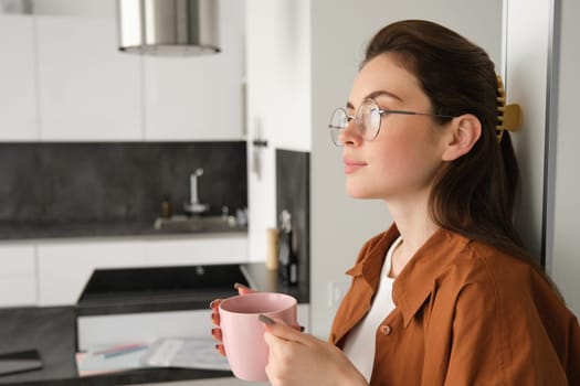 Portrait of cosy, beautiful and relaxed young woman at home, standing and leaning on wall with cup of coffee, drinking tea with thoughtful face expression.