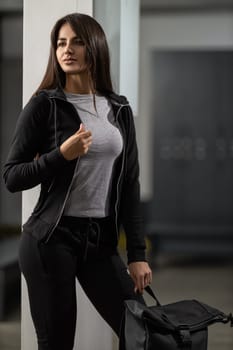 Beautiful fitness model brunette woman in a modern black tracksuit posing in the gym locker room with a sporty black backpack