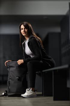 Beautiful fitness model brunette woman in a modern black tracksuit posing in the gym locker room with a sporty black backpack