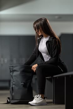 Beautiful fitness model brunette woman in a modern black tracksuit takes clothes out of black backpack in the gym locker room