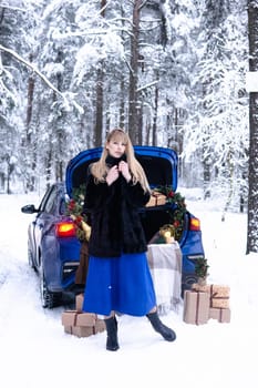 Woman in winter snowy forest in blue dress next to blue car decorated with Christmas decor. Christmas and winter holidays concept