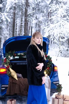 Woman in winter snowy forest in blue dress next to blue car decorated with Christmas decor. Christmas and winter holidays concept