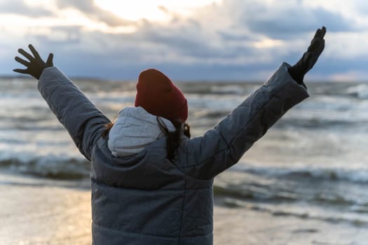 a woman stands on the seashore in the cold during a storm. The girl looks at the waves with her arms wide open