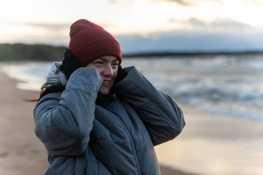 a woman stands on the seashore in the cold during a storm. The girl looks at the waves covering her ears with her hands from the wind