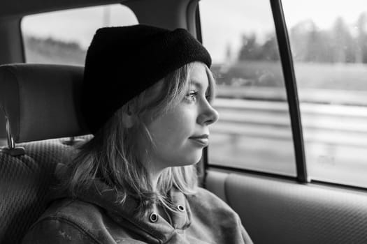 black and white photo. A pretty girl with pink hair in a black knitted hat in the back seat of a car, looking out the window, she is immersed in thoughts about her future, about opportunities. Traveling by car