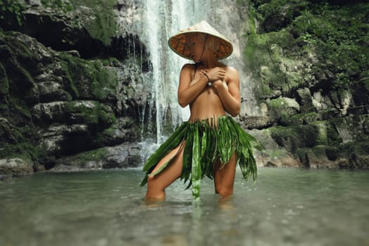 naked sexy girl in a triangular hat and a homemade cape made of leaves stands at the waterfall of a beautiful mountain river