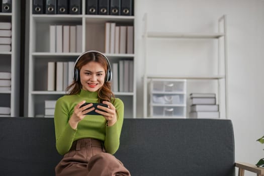 Woman wearing headphones plays video game on smartphone while relaxing on sofa in home lifestyle..