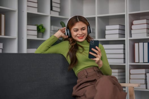 Happy Asian woman listening to music on mobile phone while sitting on sofa at home Relaxed smiling girl with headphones in the morning It's time to rest..