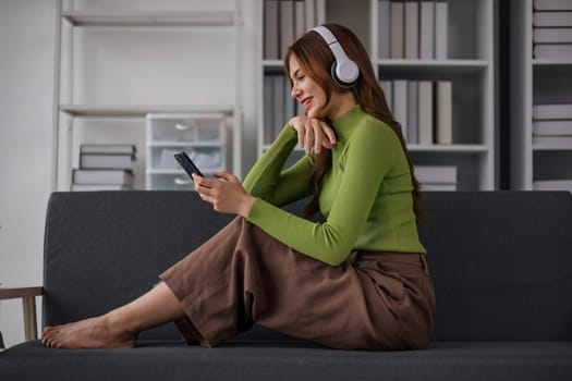 Happy Asian woman listening to music on mobile phone while sitting on sofa at home Relaxed smiling girl with headphones in the morning It's time to rest..