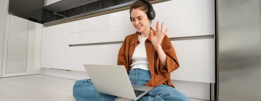 Woman sits on floor in headphones, waves hand at laptop screen, says hi on online meeting, video chats, talks with team, works from home, studies online. concept of e-learning and education.