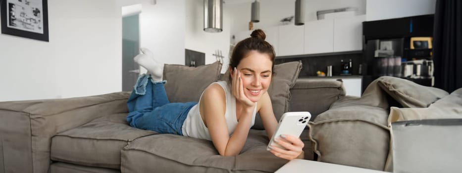 Portrait of smiling, happy young woman resting at home, resting on couch with smartphone, using mobile phone application.