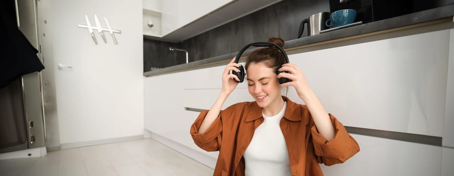 Stylish modern woman at home, sits on floor, puts on wireless headphones and smiles, listens to music, rests indoors.
