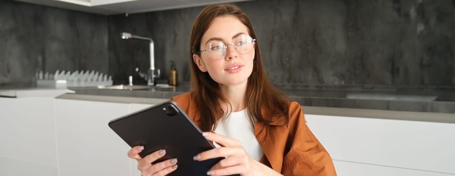 Portrait of young beautiful woman in glasses, working from home, studying online in her kitchen, holding digital tablet, online shopping.