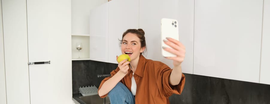 Happy woman using smartphone to take selfie, eating an apple on camera, records video for social media application, sitting on kitchen counter.